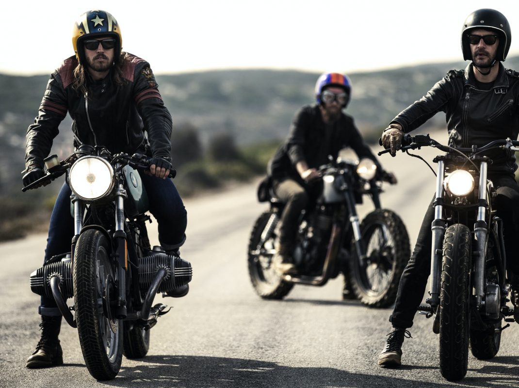 three-men-wearing-open-face-crash-helmets-and-goggles-sitting-on-cafe-racer-motorcycles-on-a-rural.jpg